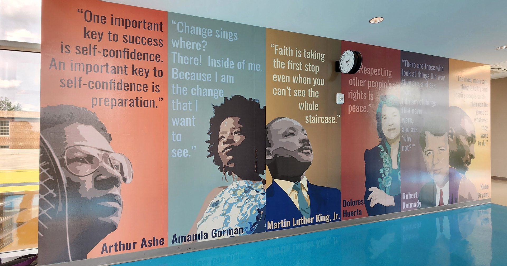 A floor-to-ceiling adhesive vinyl wall mural in a hallway. The mural features quotes and images from various individuals, each with a different color background for their rectangular space. From left to right: The first section has black text on an orange background, reading 'One important key to success is self-confidence. An important key to self-confidence is preparation.' Above an image of Arthur Ashe, with his name below in black. The second section features light blue text on a darker blue background, reading 'Change sings where? There! Inside of me. Because I am the change that I want to see.' Above an image of Amanda Gorman, with her name below in black. The third section has light yellow text on a darker goldenrod background, reading 'Faith is taking the first step even when you can't see the whole staircase.' Above an image of Martin Luther King Jr., with his name below in black. The fourth section has light pink text on a deep red background, reading 'Respecting other people's rights is peace.' Above an image of Dolores Huerta, with her name below in black. The fifth section features lilac text on a burgundy background with a quote by Robert Kennedy, above an image of Robert Kennedy with his name below in black. The final section has black text on a goldenrod background, with a quote by Kobe Bryant above an image of Kobe Bryant, with his name below in black.