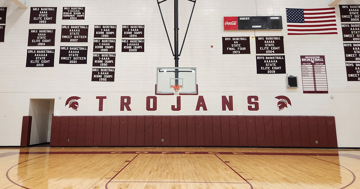 Vinyl-covered wall padding beneath adhesive vinyl lettering that reads "TROJANS," with images of Trojan helmets on both the right and left sides of the lettering. Above the padding and lettering is a basketball goal, and in front is a basketball court. Above, several black rectangular banners with white lettering display the sporting achievements of the boys' and girls' basketball teams.