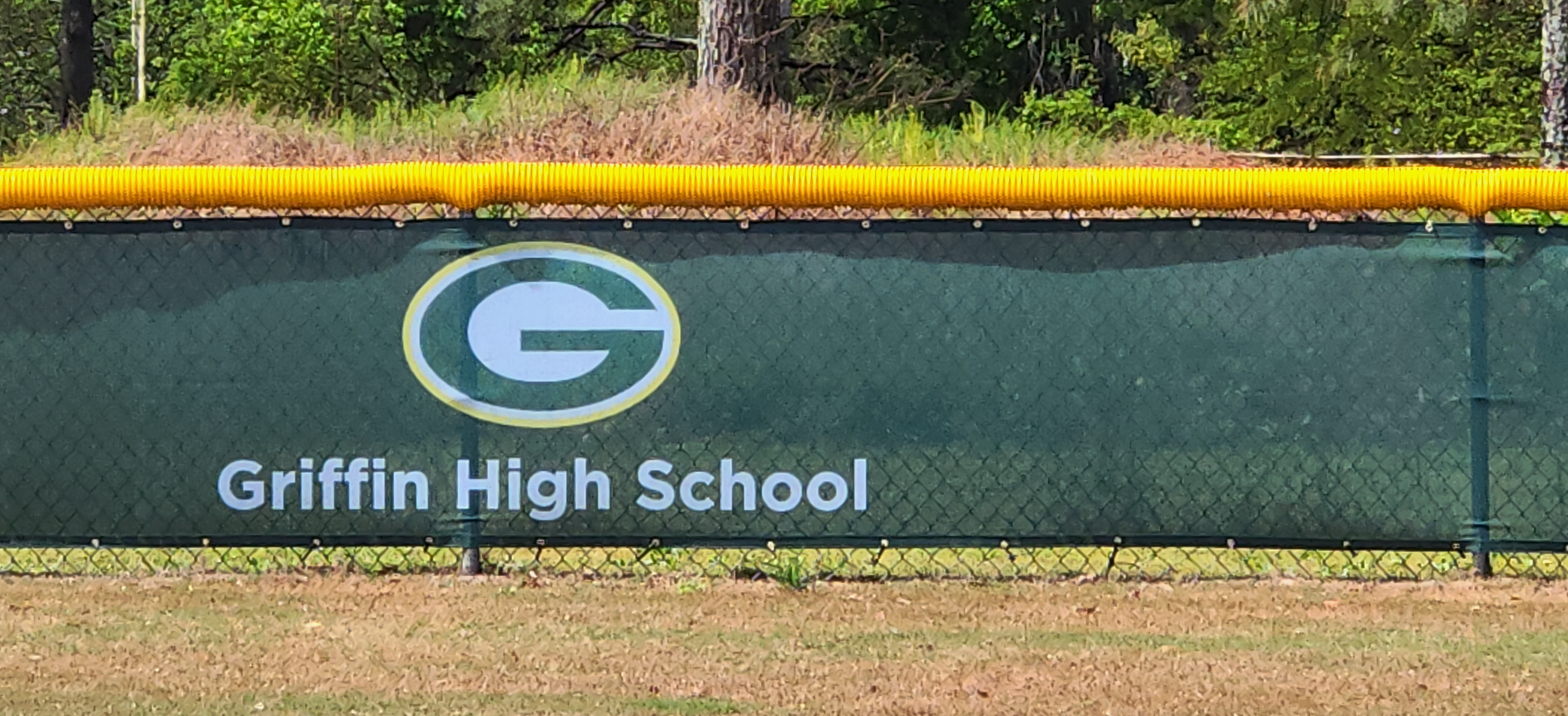 Closeup shot of dark forest green windscreen that is branded with a green, white and yellow 'G' logo reading and text reading 'Griffin High School' attached to a fence with a yellow corrugated poly cap rail top fence cap.