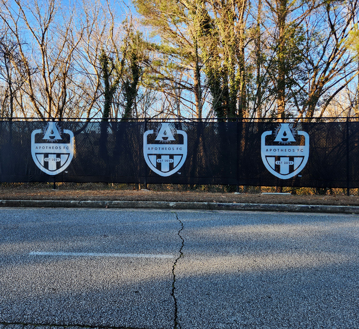 black fence windscreen on a fence along the side of a road, printed with three white logos reading 'apothes fc'
