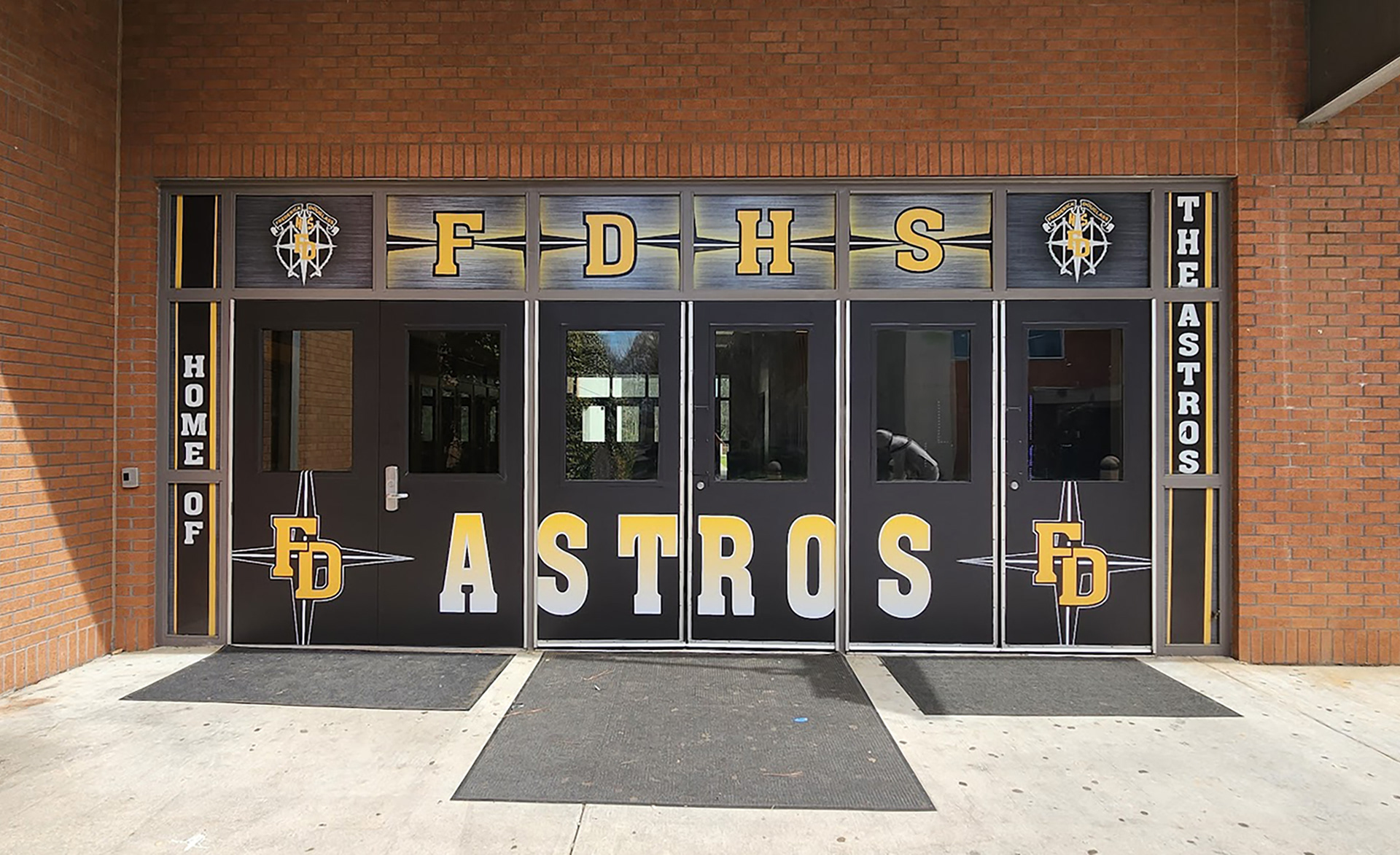 window perf reading 'FDHS' in yellow with a grey background and two 'FD' logos on either side over a set of doors. To the left of the doors full print window perf reading 'home of', to the right of the doors full print window perf reading 'the Astros', both on a black background with yellow edges. On the doors full print adhesive vinyl reading 'FD Astros FD' in yellow on a black background