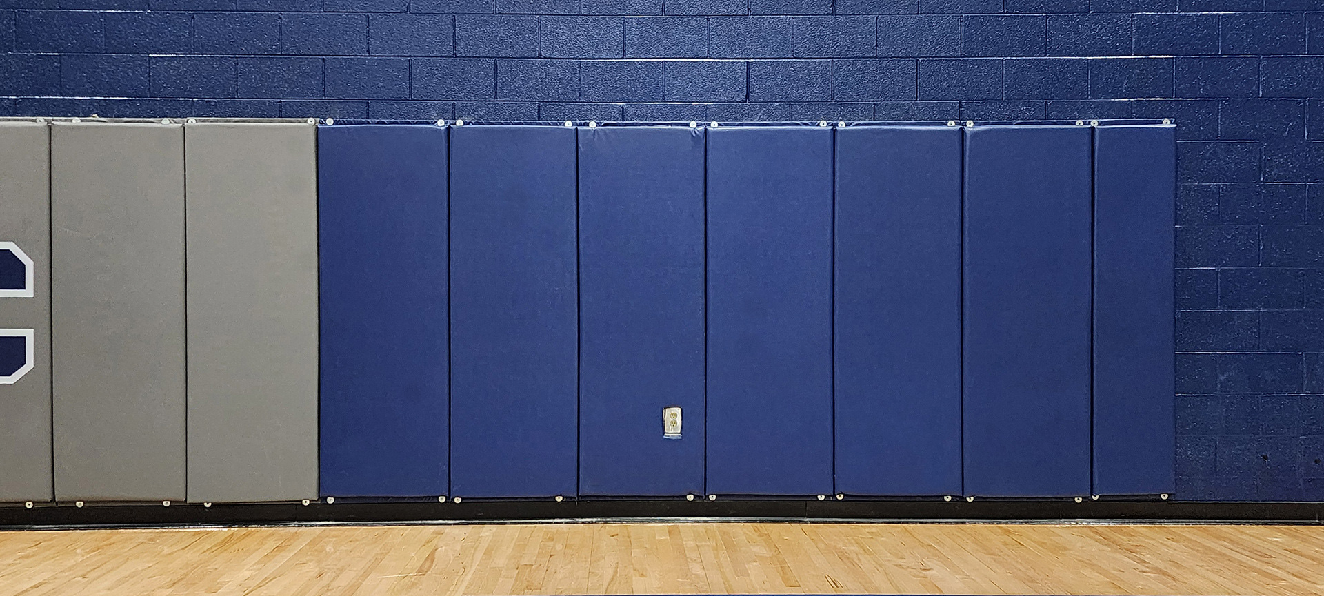 Ten vinyl-covered backstop pads attached to a painted brick wall. The three left pads are gray, with the far-left pad featuring printing. The seven right pads are blue, with one pad having a square cut-out for an outlet plug.