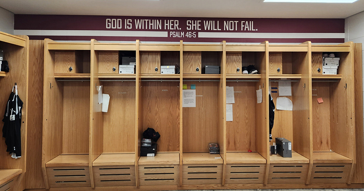 An adhesive vinyl wall decal strip running from wall to wall with a burgundy background and white lettering in a girls' locker room, above wooden lockers. . The text reads "god is within her. She will not fail." above "PSALM 46:5," with a long white line on both the right and left.