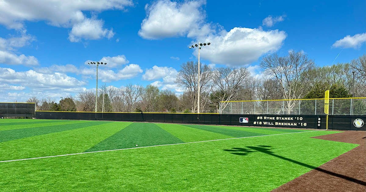 Black windscreen attached to a fence branded with an blue, red and white MLB logo next to text printed in yellowand white reading "#5 Ryne Stanek '10" and "18 Will Brennan '16". A black windscreen field marker with "300" printed in yellow and white and a black, white and yellow round logo reading "Blue Valley Tigers". Fence is topped with yellow corrugated poly cap rail top fence cap and surrounds a baseball field.