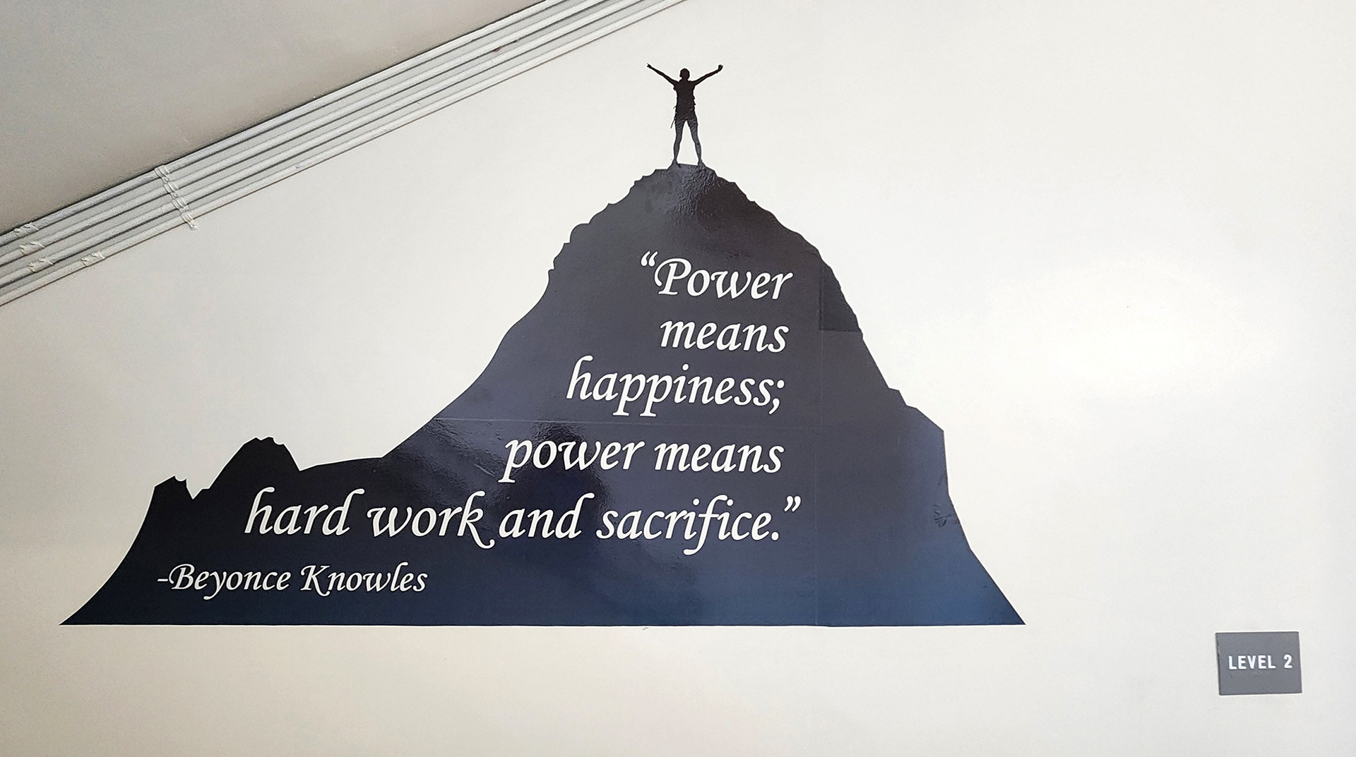 On a wall near a stairwell landing or platform, a black adhesive vinyl silhouette of a mountain with cutout lettering inside it that reads 'Power means happiness; power means hard work and sacrifice.' – Beyoncé Knowles. The silhouette also features a person with their hands raised, standing on top of the mountain. 