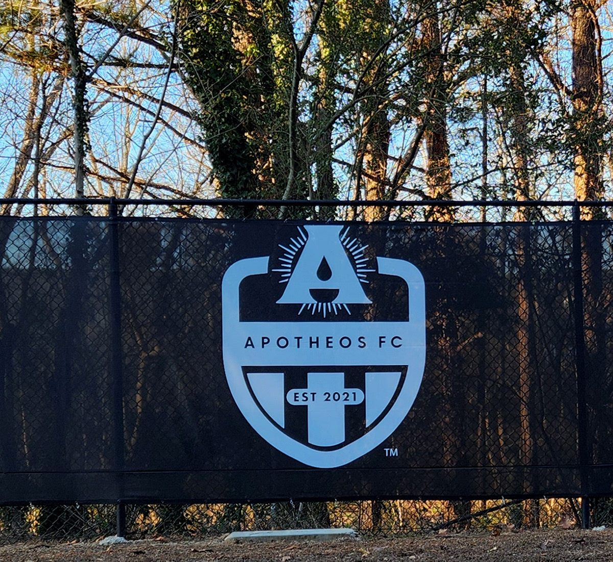 black fence windscreen on a fence along the side of a road, printed with a white logo reading 'apothes fc'