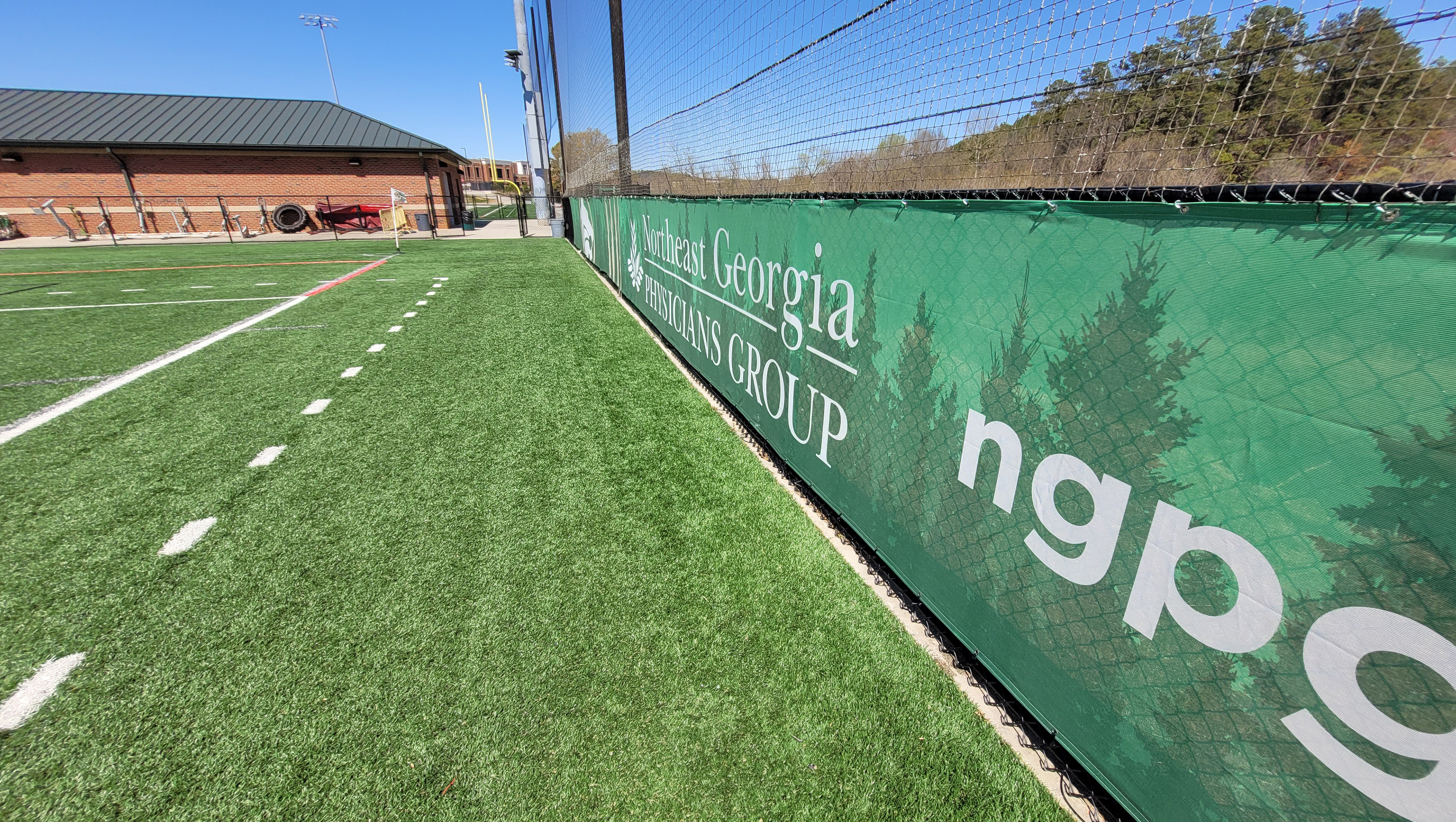 Dark evergreen tree silhouette and white lettering printed next to sponsor messages on a green fence windscreen at the end of a football field, viewed near the fence from the far right side.