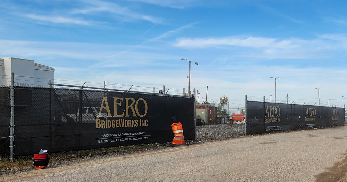A fence surrounding a business, featuring a branded black windscreen with the words 'AERO BridgeWorks Inc' printed in tan, affixed to it.
