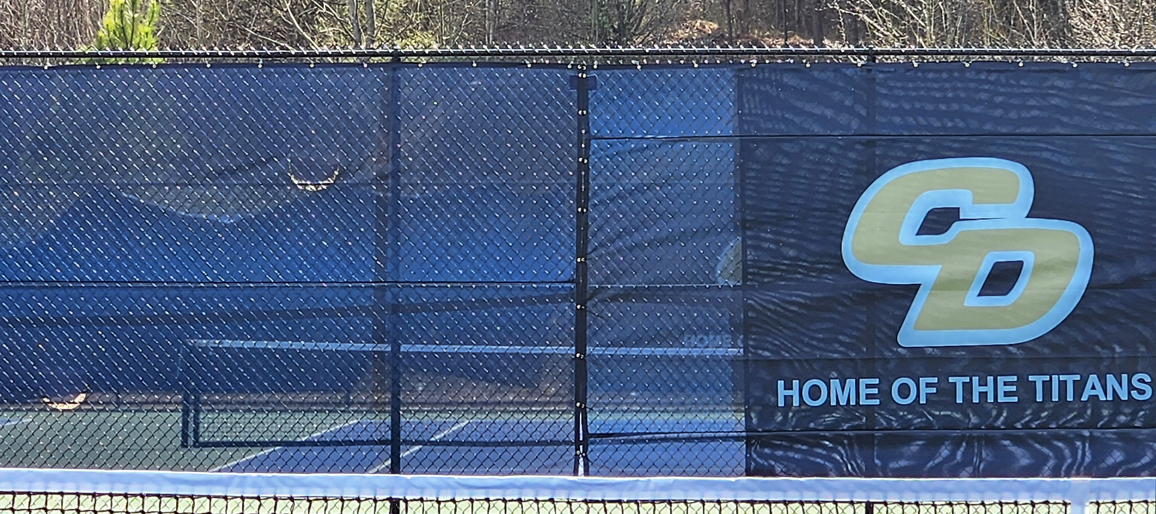 Blue fence windscreen surrounding a tennis court, featuring a yellow 'CD' monogram logo outlined in white, with the words 'Home of the Titans' displayed.