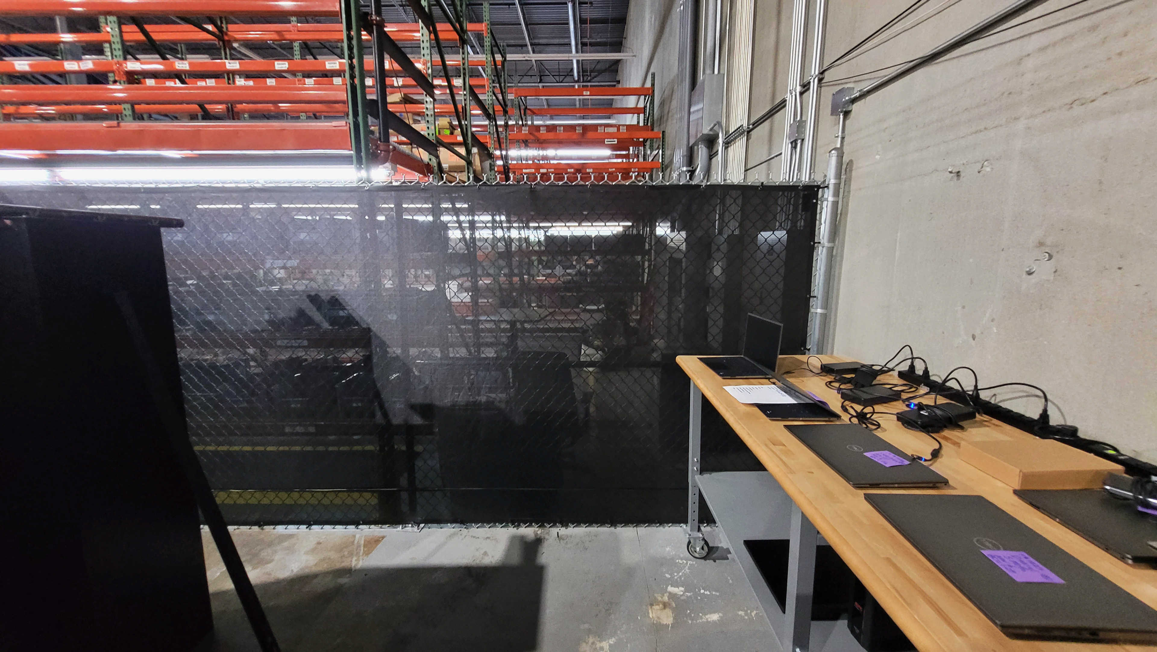 Black fence windscreen inside a warehouse, used to separate the shelving area from a computer workspace area.