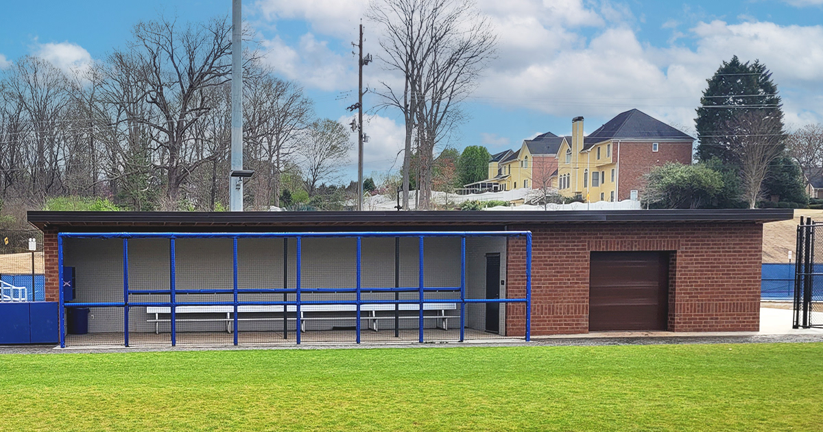 A full view of an alternate dugout, featuring round rail foam padding covered in blue sports vinyl with black sports netting in front of the barrier, viewed from the front at a distance to capture the entire dugout.