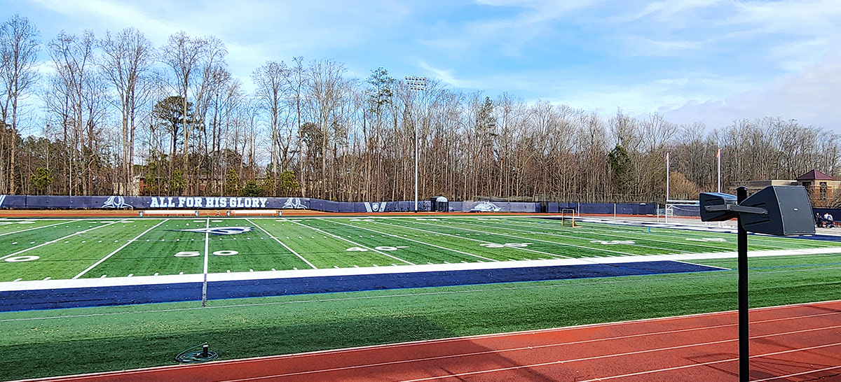 Long shot of navy blue fence windscreen panels alongside a football field, featuring from left to right in white: an outline of a knight, the words "ALL FOR HIS GLORY," another knight outline, an 'FC' monogram logo flanked by three vertical white stripes angling inward at the bottom and outward at the top, and an outline of a knight riding a horse in a jousting position.