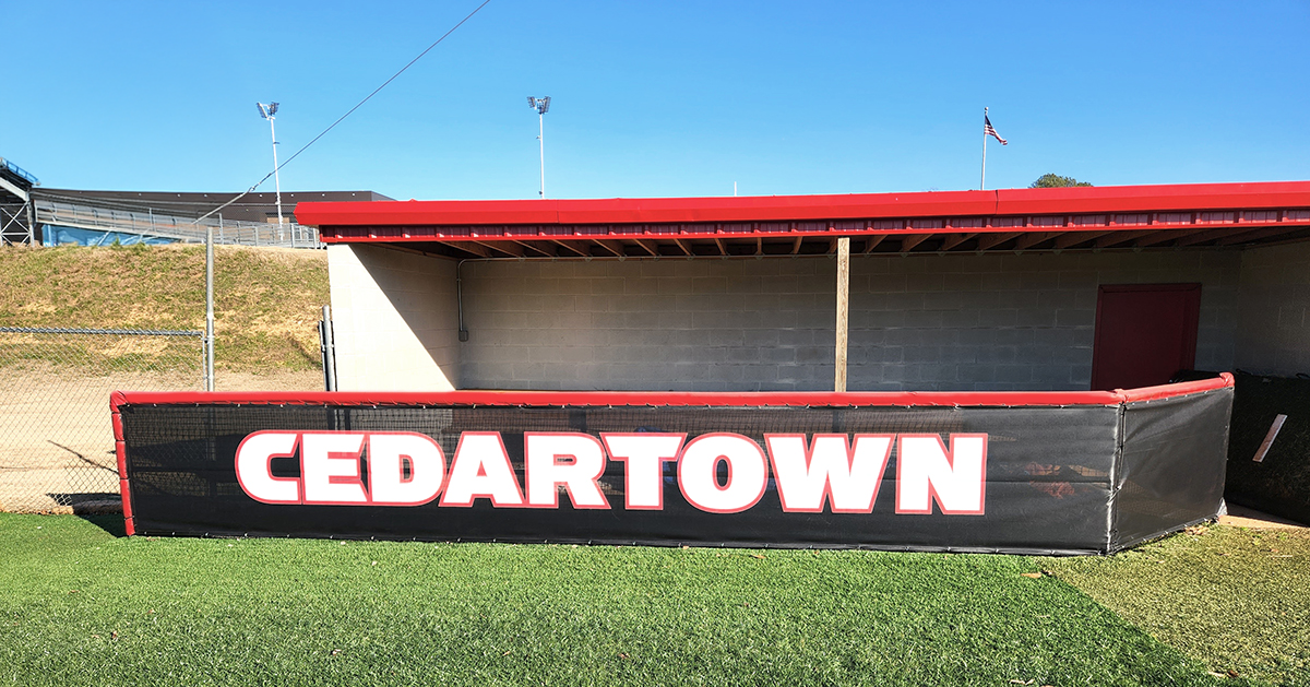Black fence windscreen for a dugout, featuring the word 'Cedartown' printed in white with a red outline. The windscreen is attached to a fence topped with red vinyl-covered round dugout rail padding.