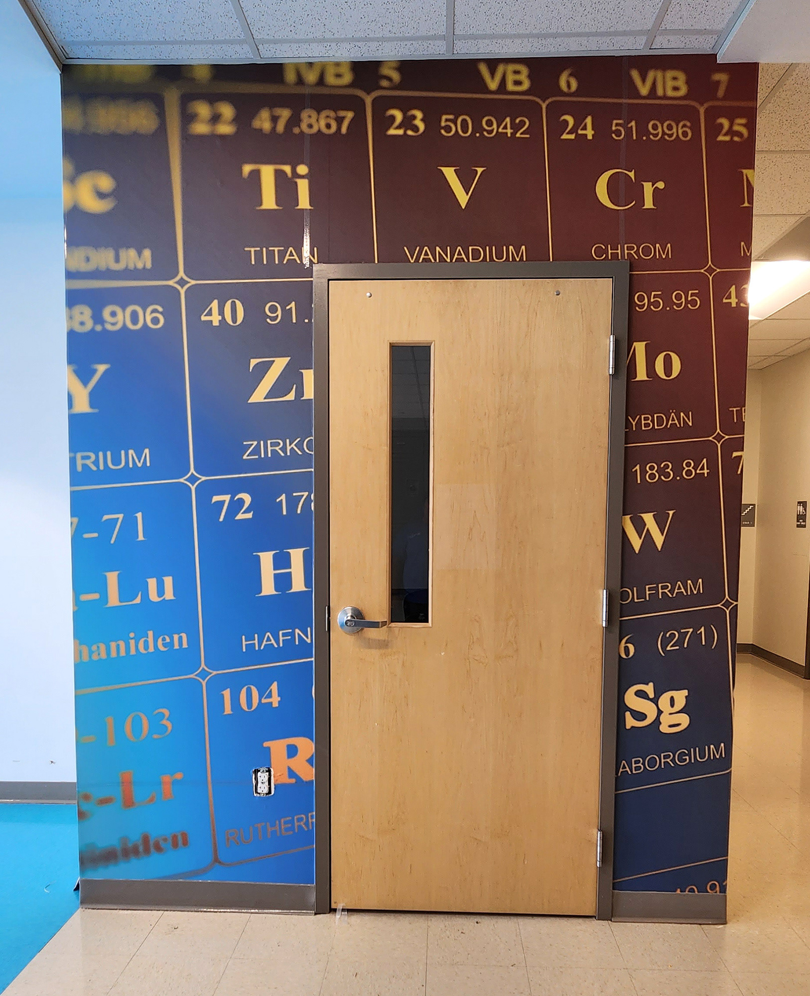 A floor-to-ceiling adhesive vinyl wall mural in a hallway with a closed door positioned in the middle and an intersecting hallway to the right. The mural features a periodic table in yellow on a gradient blue background. 