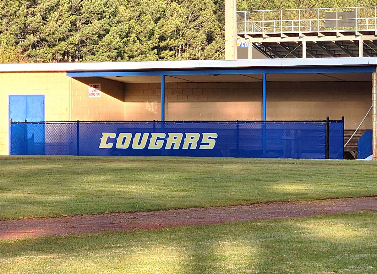 blue windscreen printed with yellow text outlined in white reading 'cougars' attached to a fence barrier surrounding a baseball dugout