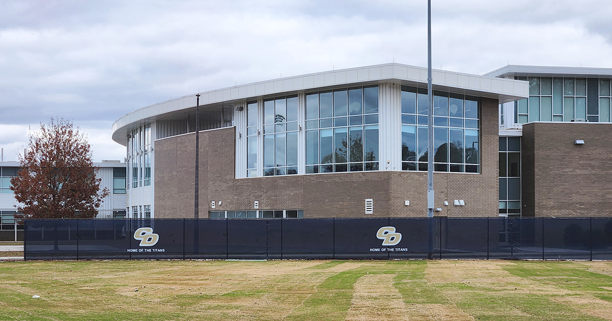Blue fence windscreen around a baseball field, featuring a repeating 'CD' monogram logo in beige outlined in white, along with the phrase 'Home of the Titans' displayed twice.
