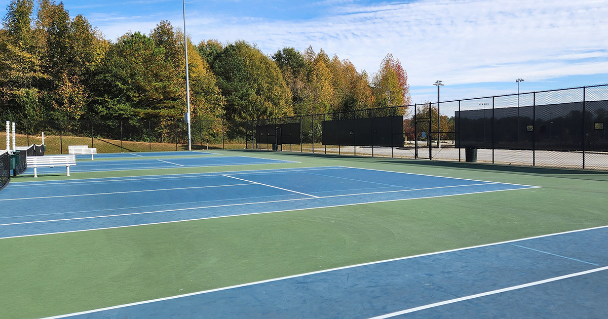 Tennis courts with a fence enclosure on the right, featuring three black windscreen panels with square vents affixed to the fence.