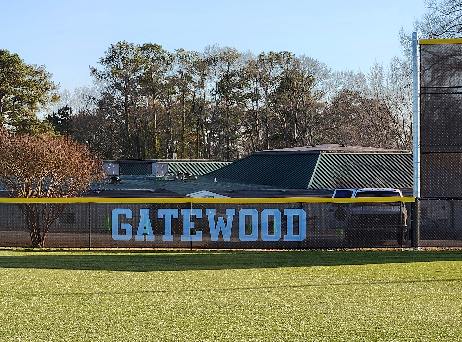 fence surrounding baseball field with black fence windscreen printed with white text reading 'gatewood' and yellow D-style safety top cap