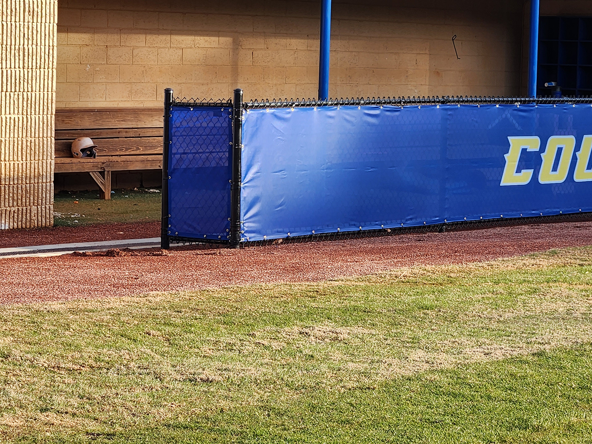 blue windscreen printed with yellow text outlined in white reading 'cougars' attached to a fence barrier surrounding a baseball dugout