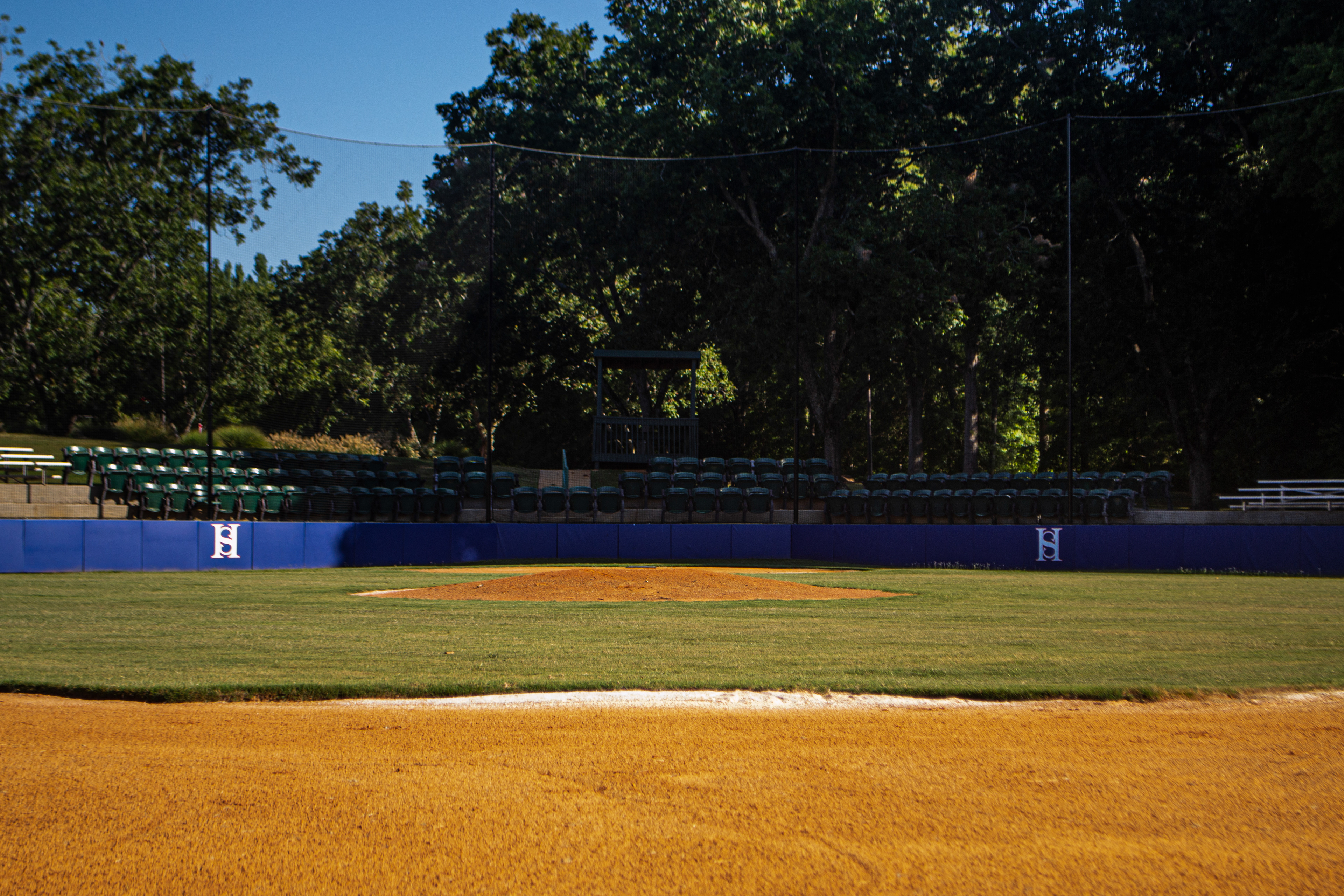 View of fencing with branded windscreen along the side of a baseball field, in front of a spectator seating section, from the viewpoint of someone standing in the infield. Raised vertically from the fencing is black sports sideline netting.