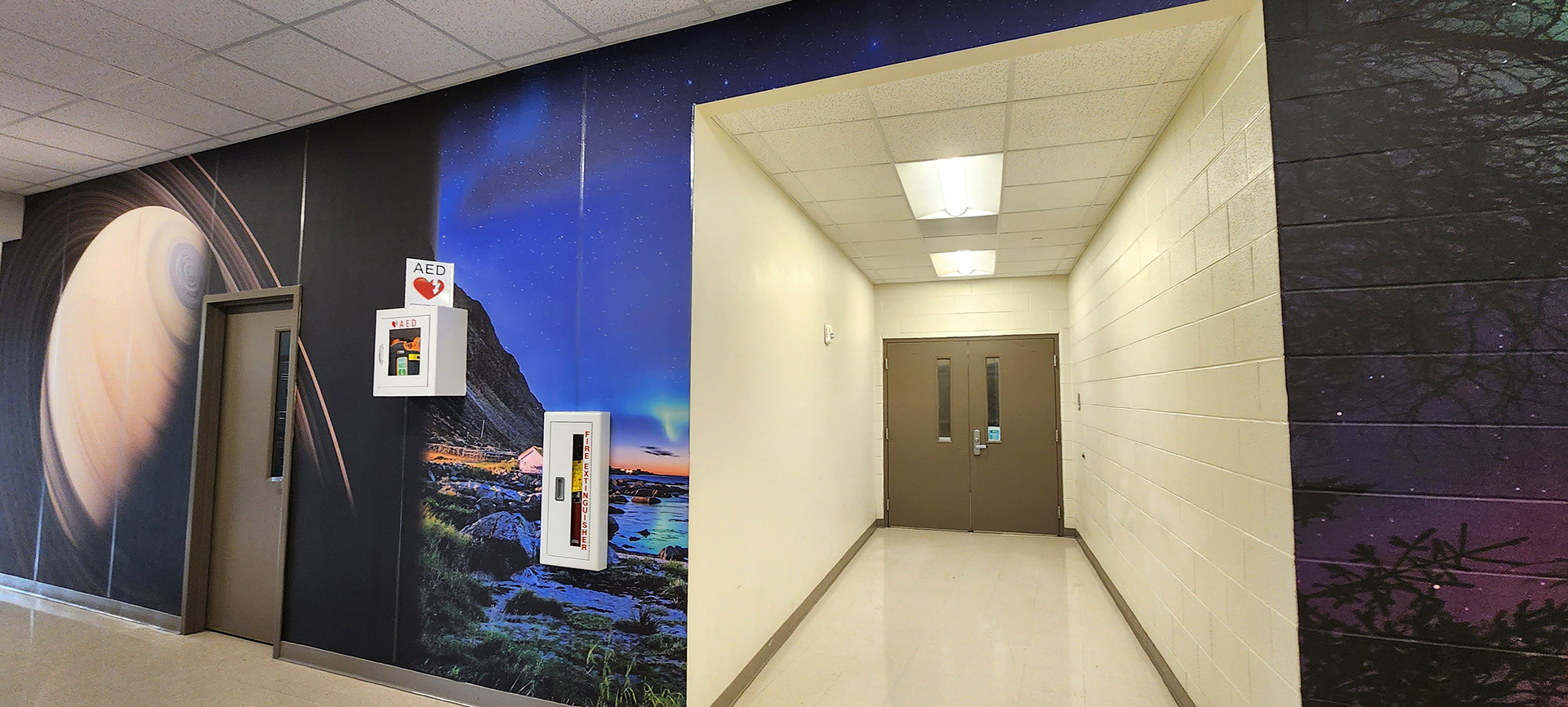 A view of the same floor-to-ceiling adhesive vinyl wall mural in a hallway from the opposite angle. The mural features, from left to right, an image of saturn, an image of the northern lights over a rocky gulf, and a sky at night over trees