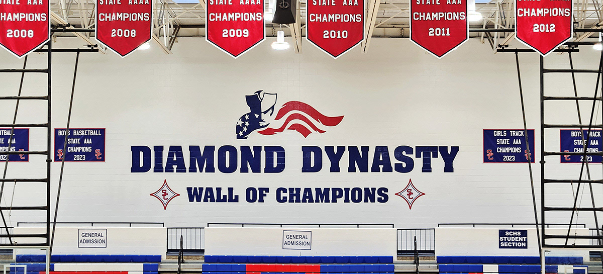 A full-print custom-cut adhesive vinyl decal in navy blue and red featuring an image of a patriot and a flag, displayed on a brick wall inside a school gymnasium. The decal is positioned above the words 'Diamond Dynasty' and 'Wall of Champions,' both in navy blue adhesive vinyl, with two red, four-point SC logo stars on either side.