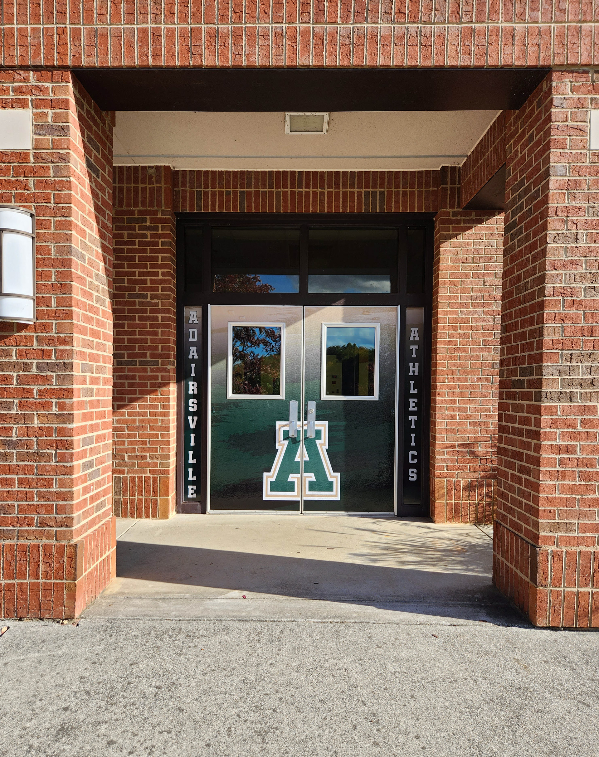 Double door entrance to a school with full print dual door wraps, creating a singular image with a gradient background and an outlined 'A' monogram in front. On either side of the doors are vertical windows with full print window perf; the left window displays the text 'ADAIRSVILLE' and the right window shows the text 'ATHLETICS'