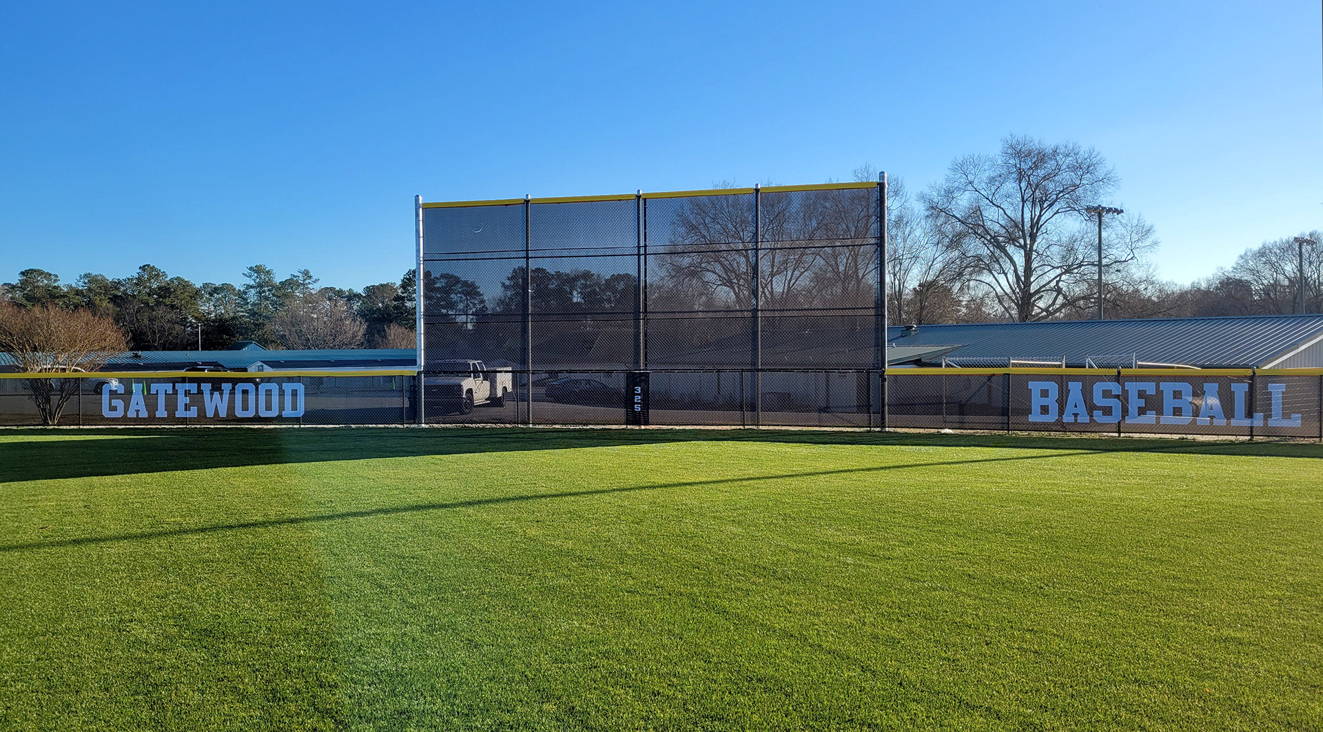 fence surrounding baseball field with black fence windscreen printed with white text reading 'gatewood baseball' and yellow D-style safety top cap
