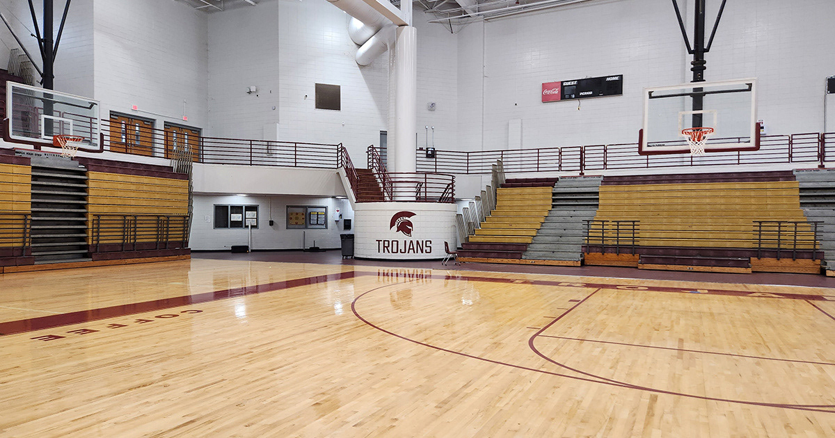Adhesive vinyl lettering reading "TROJANS," with an image of a Trojan helmet above it, located on the outside of a brick stair landing at the far end of an indoor basketball court, between the bleachers and near the concessions.