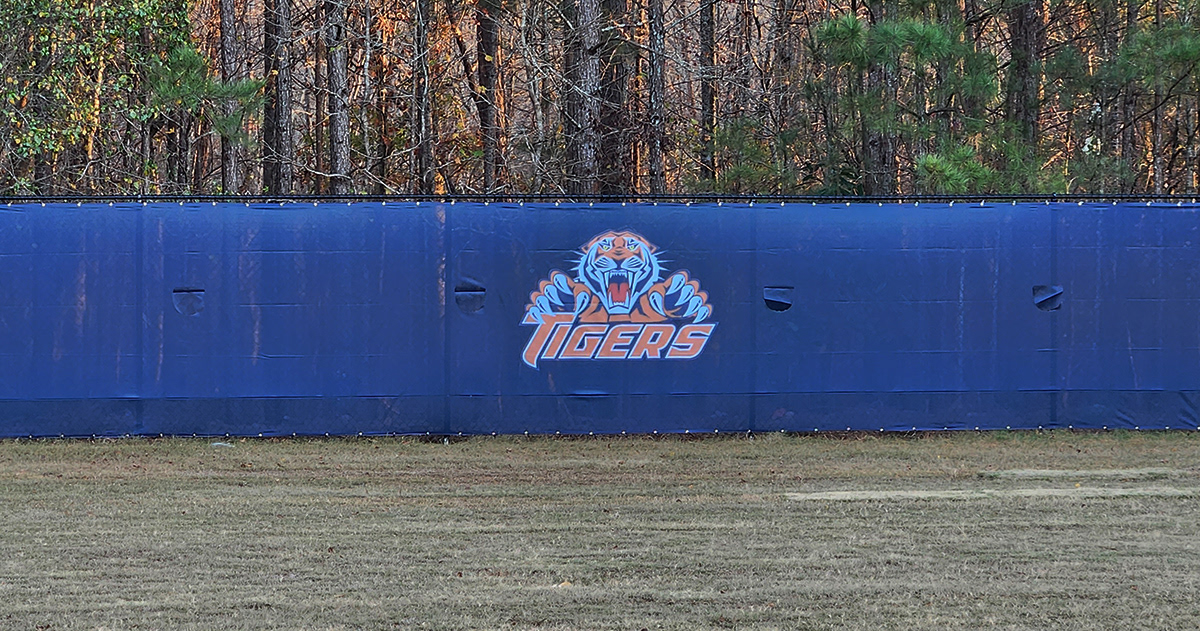 A closeup of blue fence windscreen around a baseball field featuring a tiger image above the word 'Tigers' printed in orange with a white outline