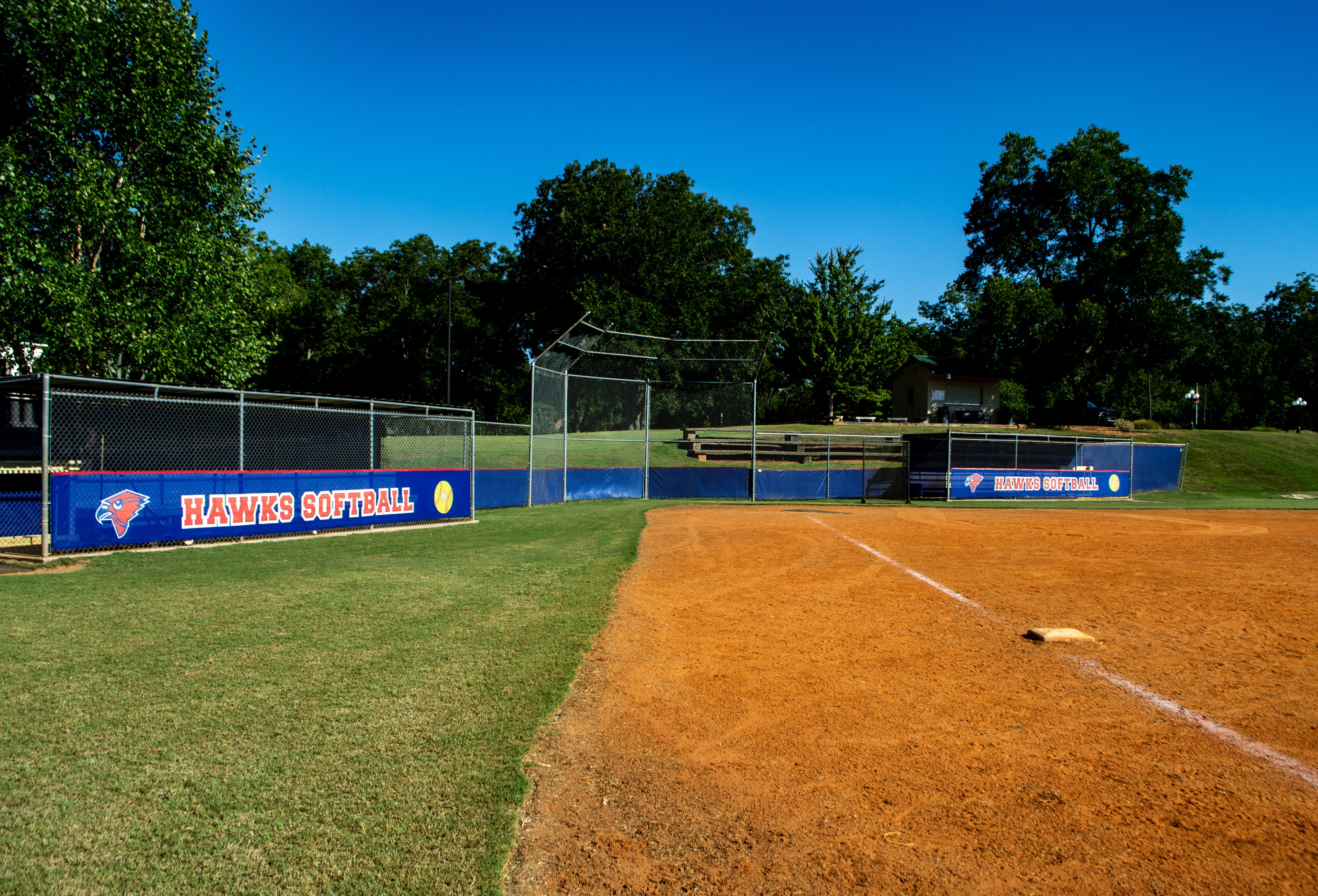 View from inside a baseball field looking at fencing around the home plate area. The bottom half of the fence has a branded windscreen with red binding. In front of the dugouts, there are windscreen panels featuring a red cardinal head logo outlined in blue, with a blue beak and white outline, and the text 'HAWKS SOFTBALL' printed in red and white. A yellow softball logo with red laces outlined in red is also visible. Behind home plate, there is a plain blue fence windscreen with red binding but no branding. Wooden bleachers and trees can be seen in the background