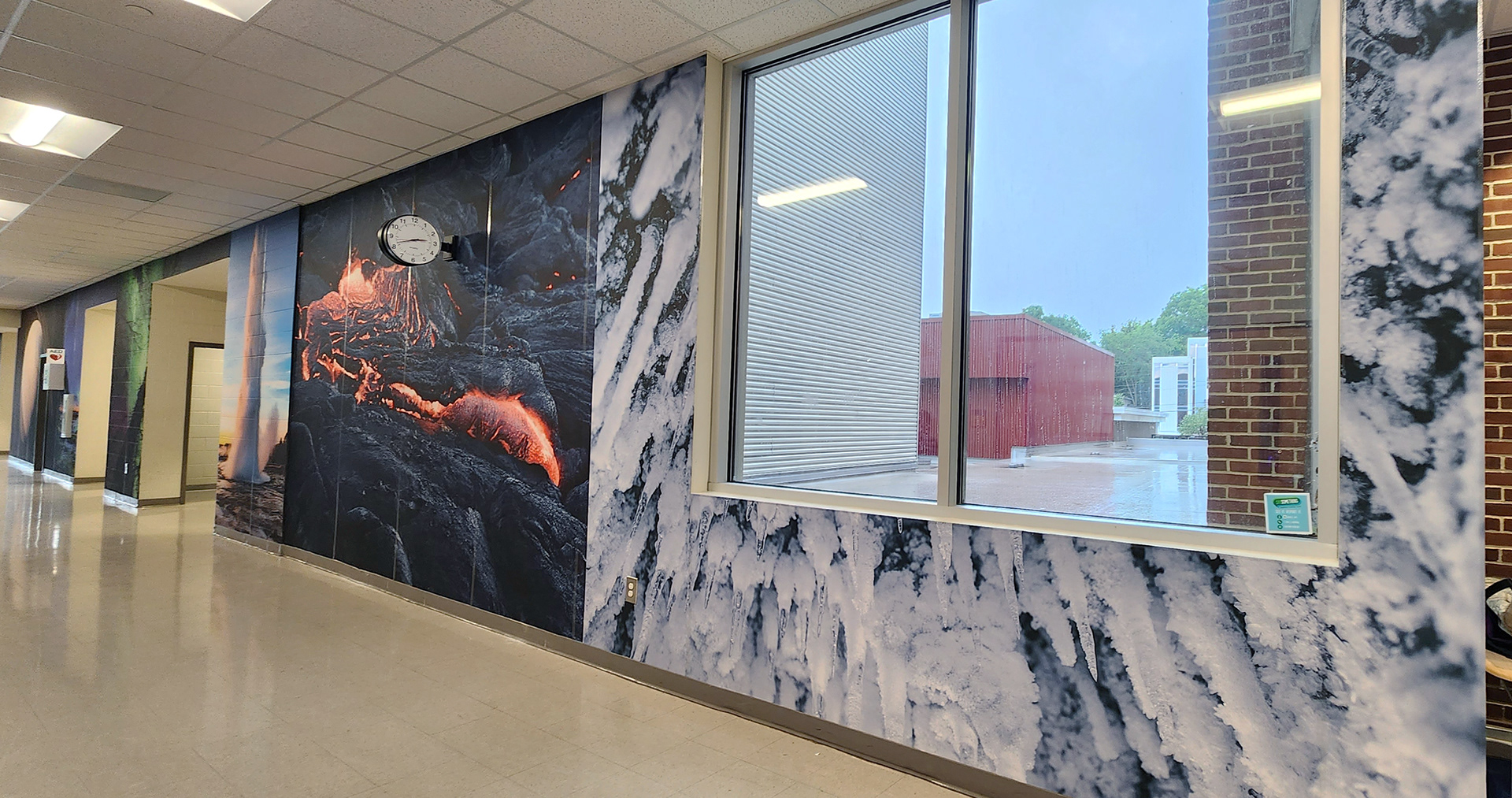 A floor-to-ceiling adhesive vinyl wall mural in a hallway. The mural features, from right to left, a glacier, lava among a volcanic cliffside, a geyser, and beyond that, the images are too distant to discern clearly. 