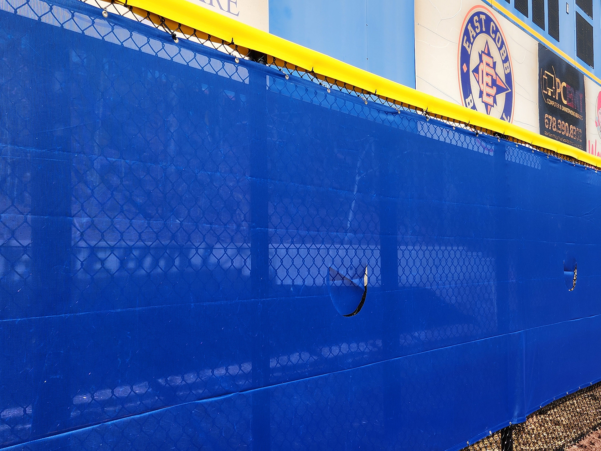 blue windscreen attached to a fence barrier topped with yellow fence guard rail top tear drop fence cap underneath a scoreboard on the outter edge of a baseball field