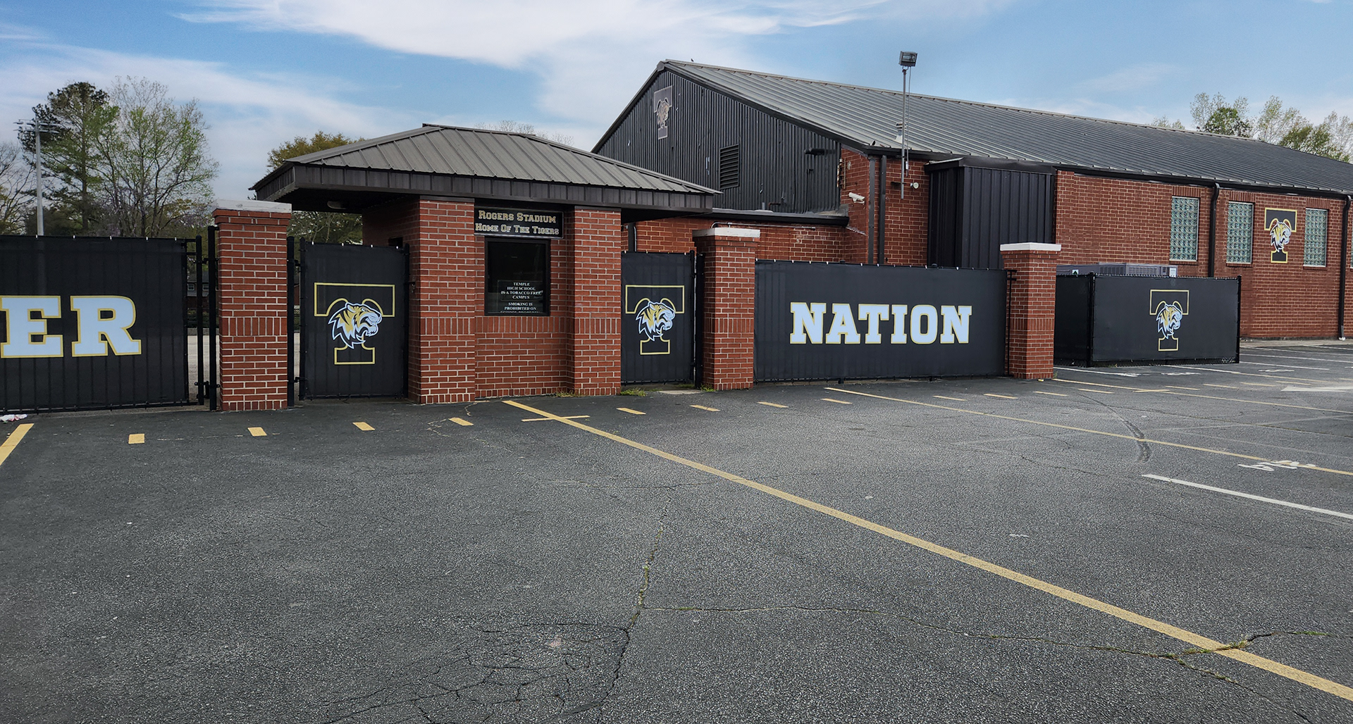 A wide shot of black windscreens branded with the letters "ER" two 'tiger nation' logos, and the words "nation" attched to fencing near a parking lot and small brick structures. Black windscreen branded with the 'tiger nation' logo attched to fencing surrounding an large AC unit near a parking lot and a large brick building branded with an adhesive vinyl 'tiger nation' logo.