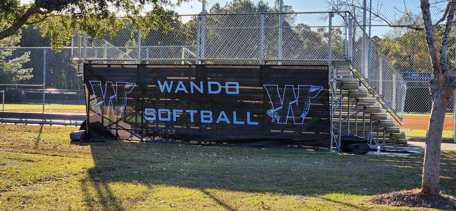 A black windscreen on the backside of metal bleachers near a baseball field. The windscreen features the text 'Wando Baseball' in white, with the 'W' logos on both sides. Each 'W' logo consists of a large black 'W' outlined in burgundy and white, with an arrow weaved through the letter.