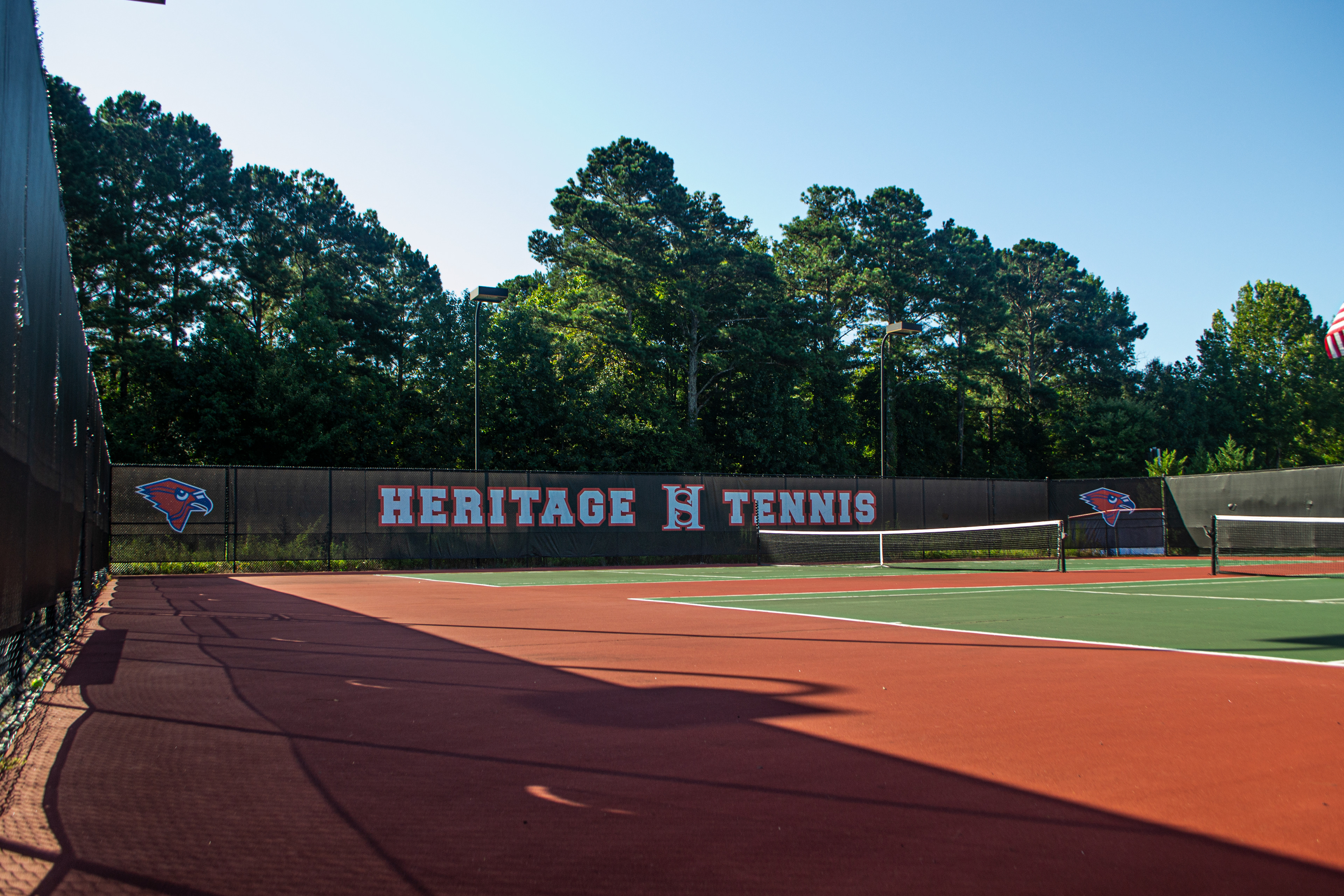 View from inside a tennis court looking at black branded windscreen on a fence at the far end of the court. The windscreen is printed from left to right with a red cardinal head with a black beak, outlined in blue, and again outlined in white. Several feet away are the words 'HERITAGE HS TENNIS,' with the 'HS' as a monogram logo, showing the two letters as one, printed in white and outlined in red. Further along, another identical cardinal head logo is printed, similar to the first
