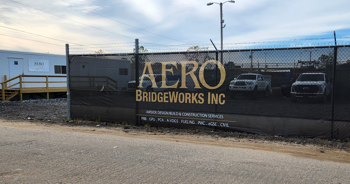 A fence surrounding a business, featuring a branded black windscreen with the words 'AERO BridgeWorks Inc' printed in tan, affixed to it.