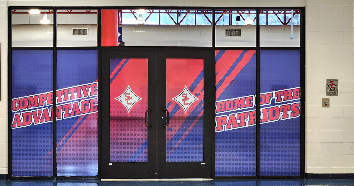 A red, white, and blue window perforation featuring the SC logo, with the text 'Competitive Advantage' and 'Home of the Patriots' displayed on three glass doors inside a school
