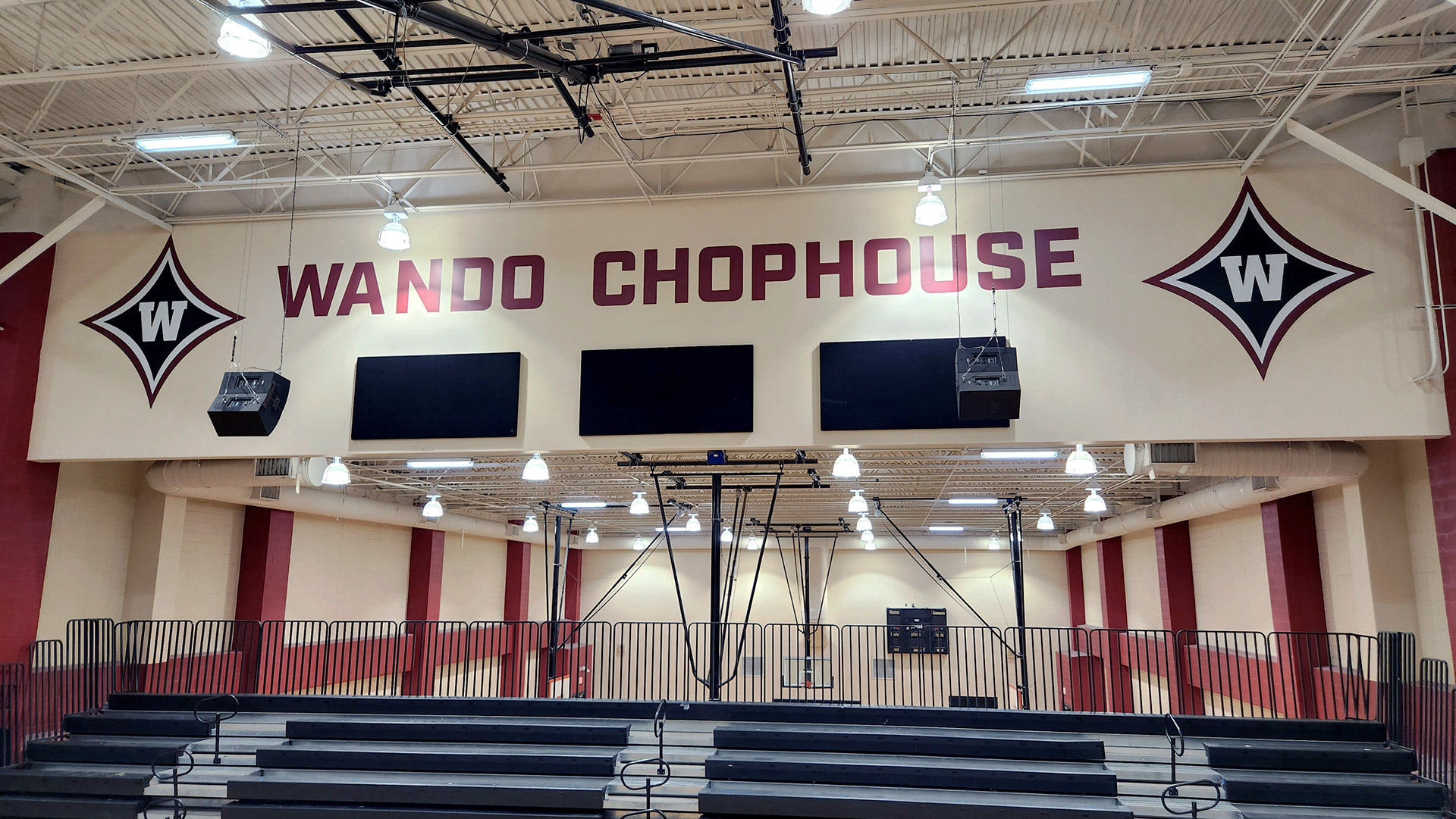 Two large adhesive vinyl navy four-point star logos, outlined in white and then red, with a 'W' in the center of each star. Between the stars, large adhesive vinyl cutout lettering in burgundy reading 'Wando Chophouse.' The logos and lettering are displayed on the upper part of gym walls near the ceiling above the bleachers.
