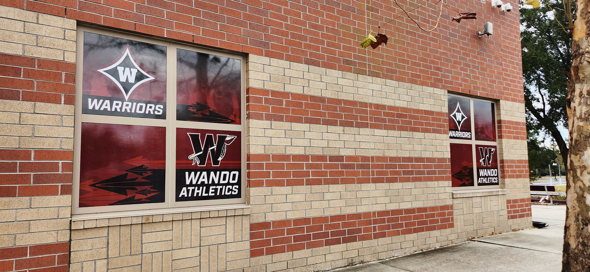 Two identical window perforations on different windows, each with a dark red translucent background. In the top left corner of each window, there is a navy four-point star logo, outlined in white and red, with a 'W' in the center and the word 'Warriors' printed in white beneath the star. In the bottom right corner, a large black 'W' logo, outlined in burgundy and white, with an arrow weaved through the letter.