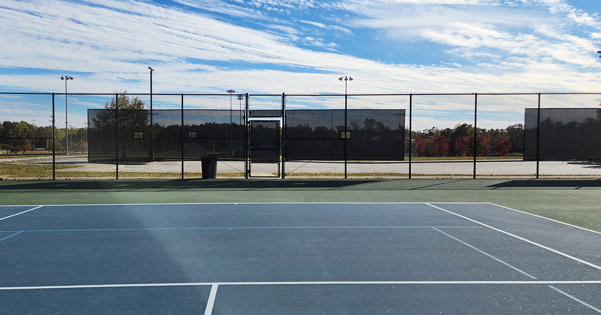 Tennis courts with a fence enclosure, affixed with black windscreen panels featuring square vents, located on, above, and to the left and right of the entrance door, with another on the far right.