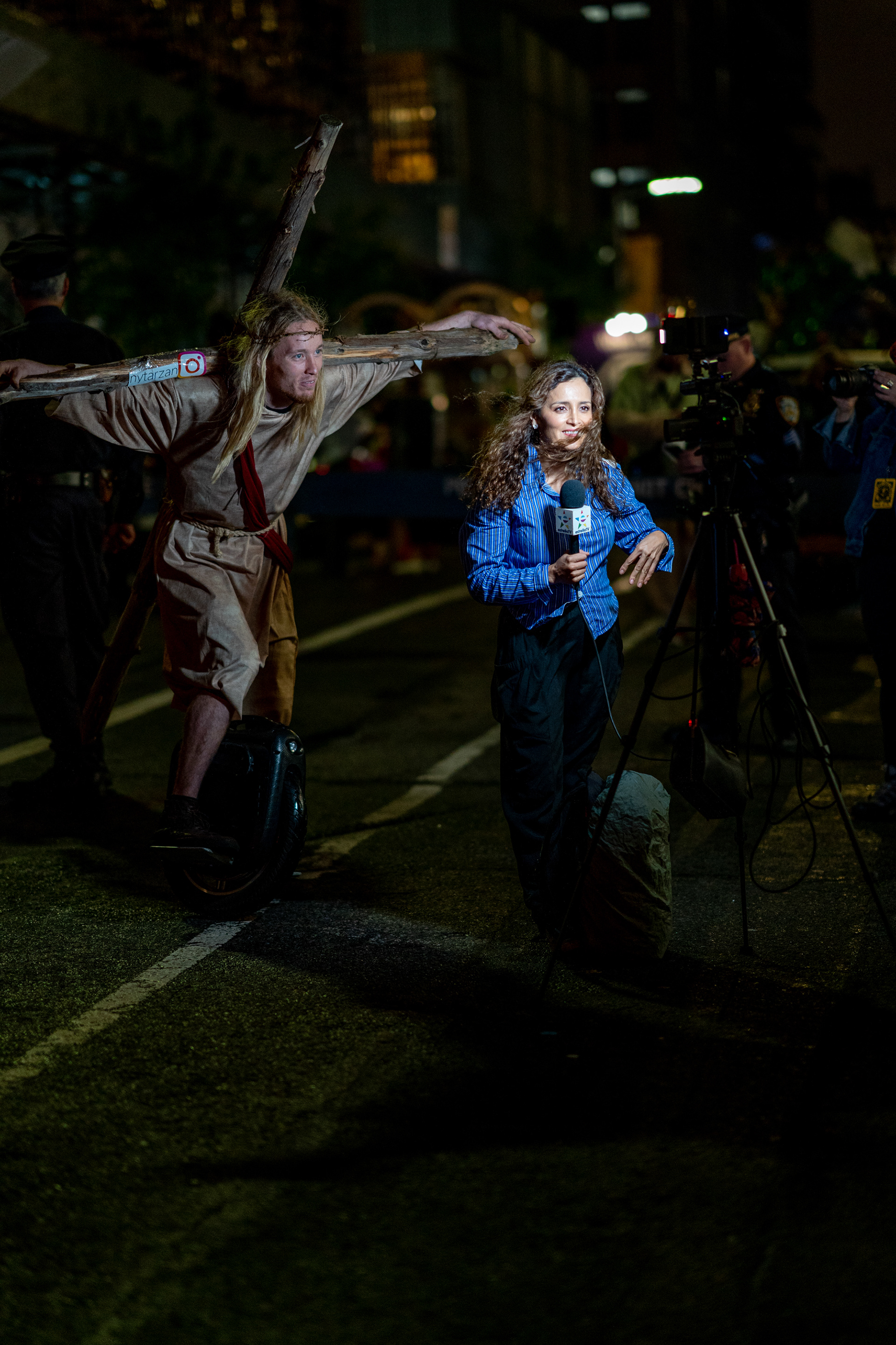 Village Halloween Parade — New York, NY. October 31st, 2019. Jesus on a Segway lurks in the background of an Estrella TV broadcast before the parade began.