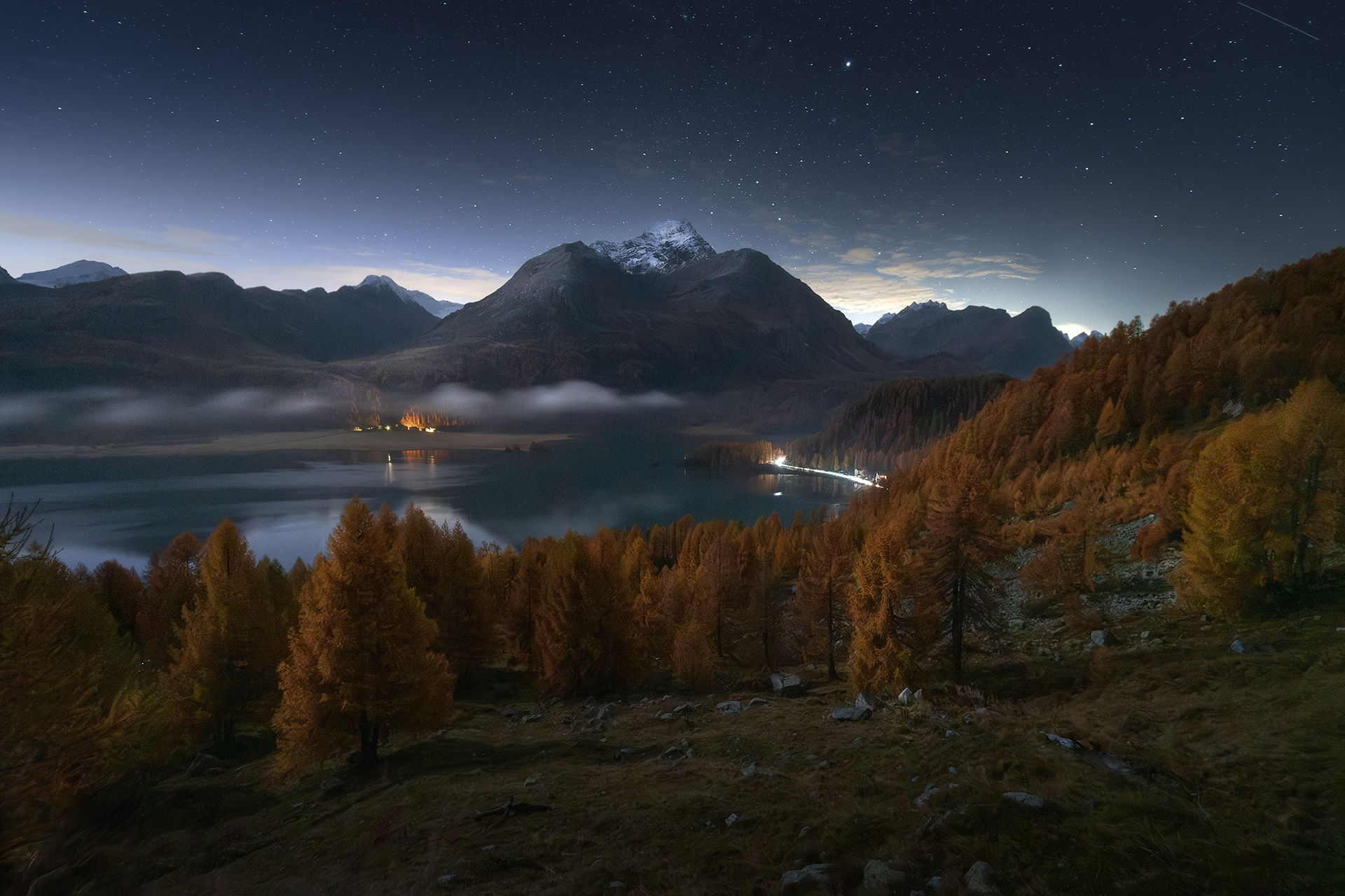 Un’atmosfera magica avvolge il lago Sils durante una serata autunnale in Engadina, Svizzera. Le cime innevate, i colori caldi dei larici e il cielo stellato creano un paesaggio surreale e incantato, perfetto per chi ama la fotografia paesaggistica e notturna.