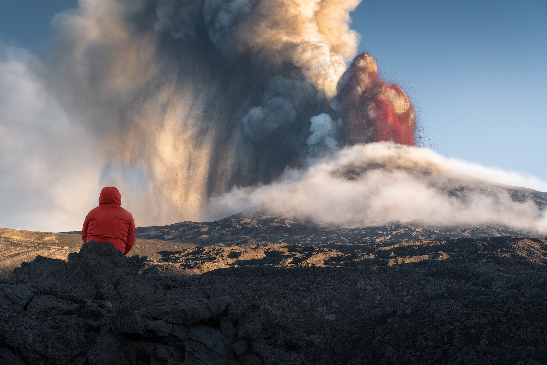 Spettatore davanti all’esplosione vulcanica Una figura solitaria in giacca rossa osserva l’Etna in eruzione, con colonne di fumo e lava che si innalzano nel cielo. Un’immagine simbolo della piccolezza dell’uomo di fronte alla potenza della natura.