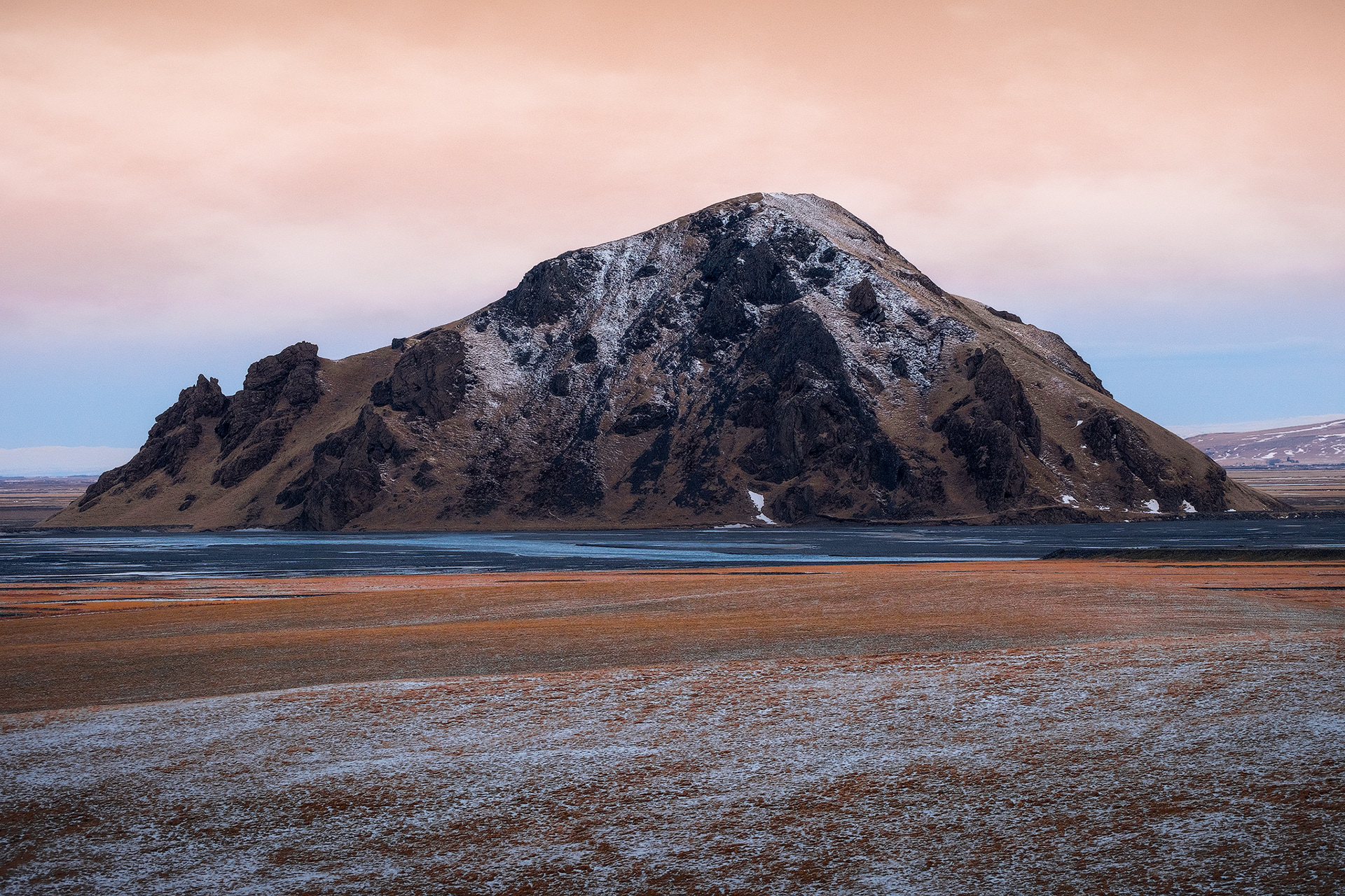 Montagna vulcanica innevata – Paesaggio remoto dell’entroterra islandese, con una montagna solitaria che emerge tra la neve e i toni caldi del cielo.