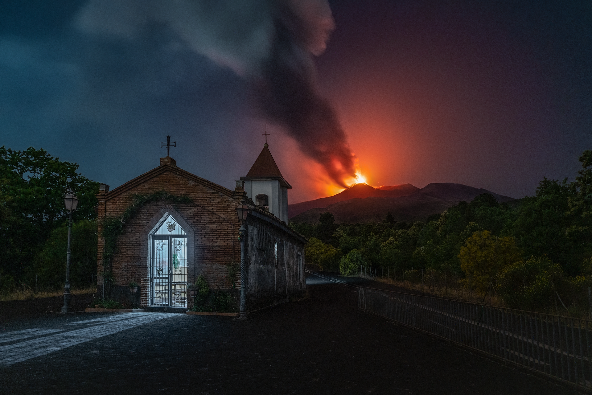 Chiesa e vulcano in eruzione Una piccola chiesa ai piedi del vulcano fa da contrasto alla furia dell’Etna in piena eruzione. Il cielo è illuminato dal bagliore della lava incandescente, creando un’atmosfera surreale e potente.