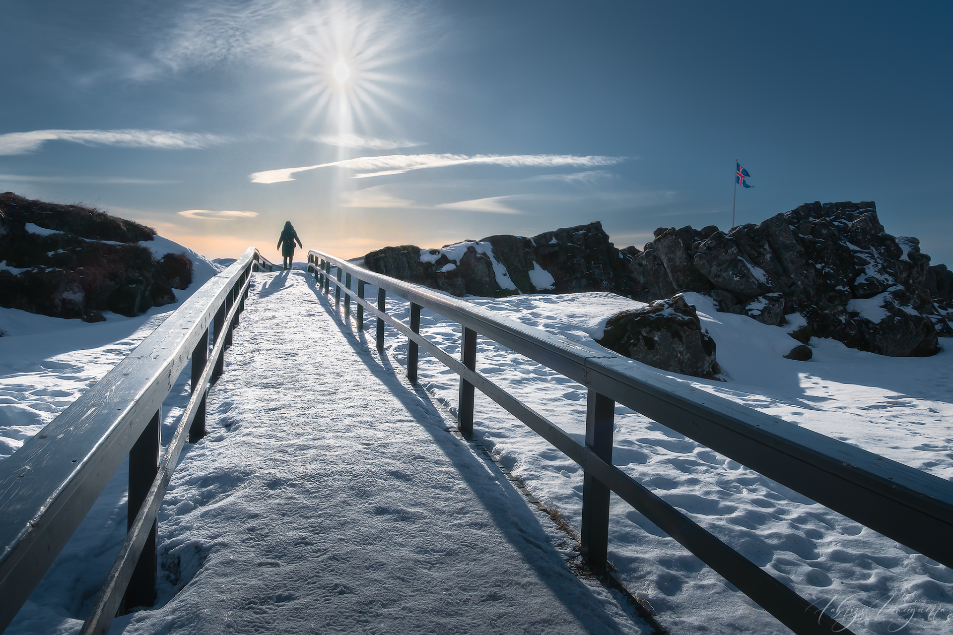 Passerella innevata verso il sole – Un viaggiatore solitario cammina su una passerella di legno, circondato dalla neve, con il sole basso che crea un effetto drammatico.