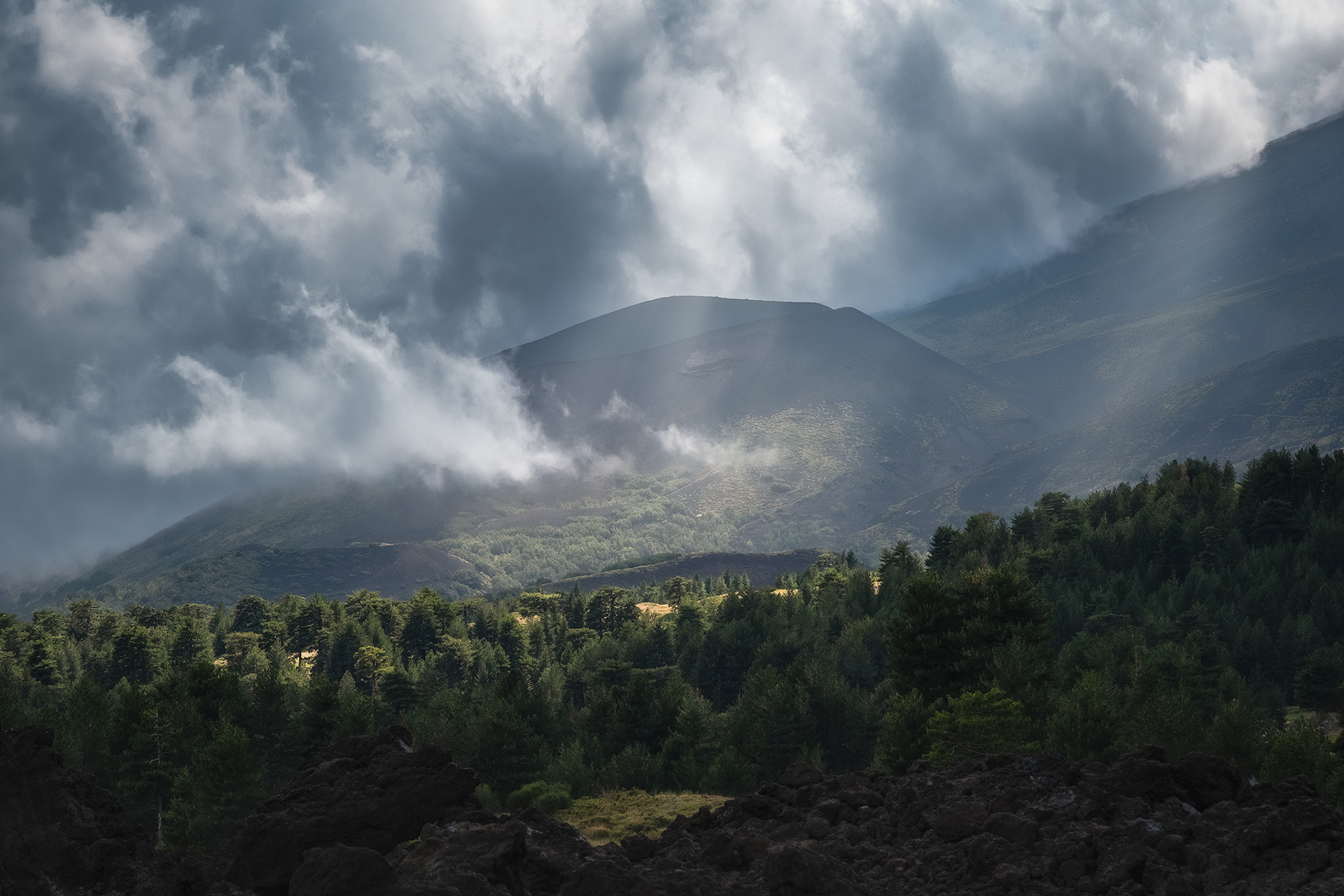 Colate laviche e foreste sotto un cielo tempestoso Una veduta del paesaggio etneo mostra il contrasto tra la vegetazione e le distese di lava solidificata, con nuvole cariche di pioggia che creano un’atmosfera dinamica e drammatica.