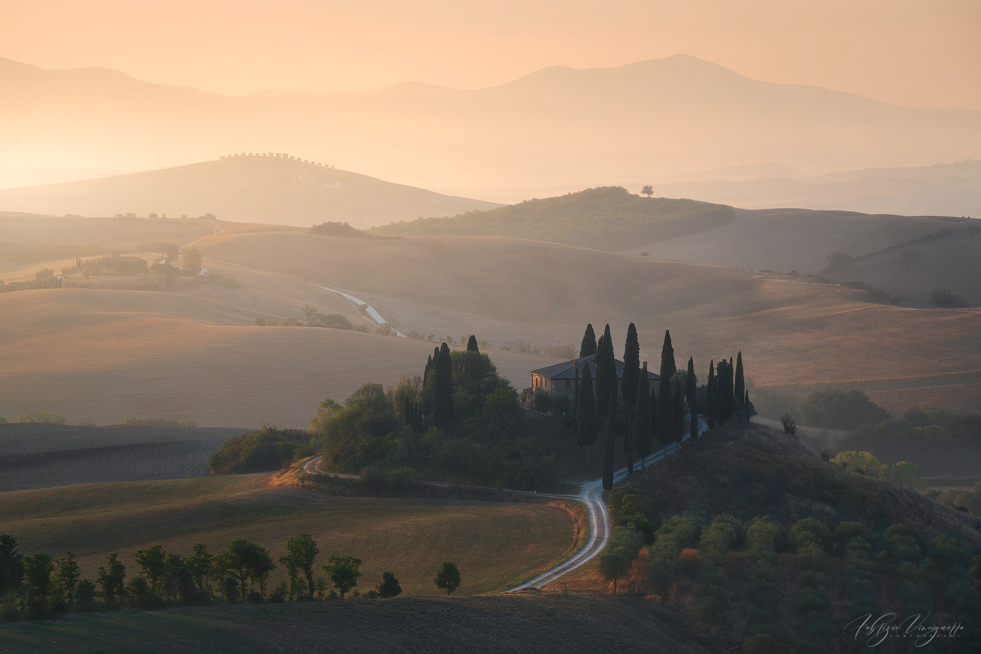 Le morbide colline della Val d’Orcia, in Toscana, emergono dalla nebbia dorata dell’alba. Un piccolo casale circondato da cipressi si trova al centro della scena, incorniciato da una luce soffusa che rende il paesaggio incantevole.