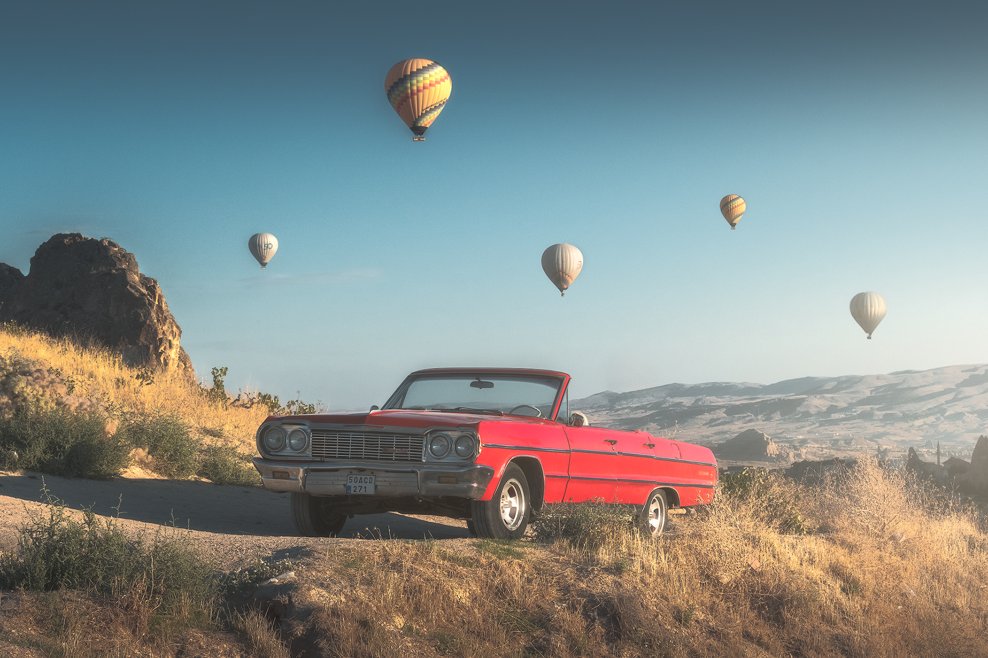 Una decappottabile d’epoca rossa è parcheggiata su una strada sterrata, immersa nel paesaggio dorato della Cappadocia. Sullo sfondo, le famose mongolfiere volano nel cielo turchese, creando un contrasto suggestivo tra il fascino vintage dell’auto e l’atmosfera onirica della regione. La luce calda e il cielo limpido esaltano la bellezza senza tempo di questo scenario unico.